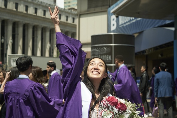 Lily in a purple graduation gown, arm raised, holding roses